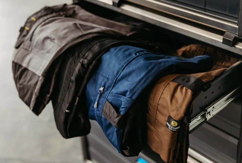 Mehrere Arbeitshosen in Kurzgröße, darunter blaue, braune und graue Modelle, liegen in einer Schublade.