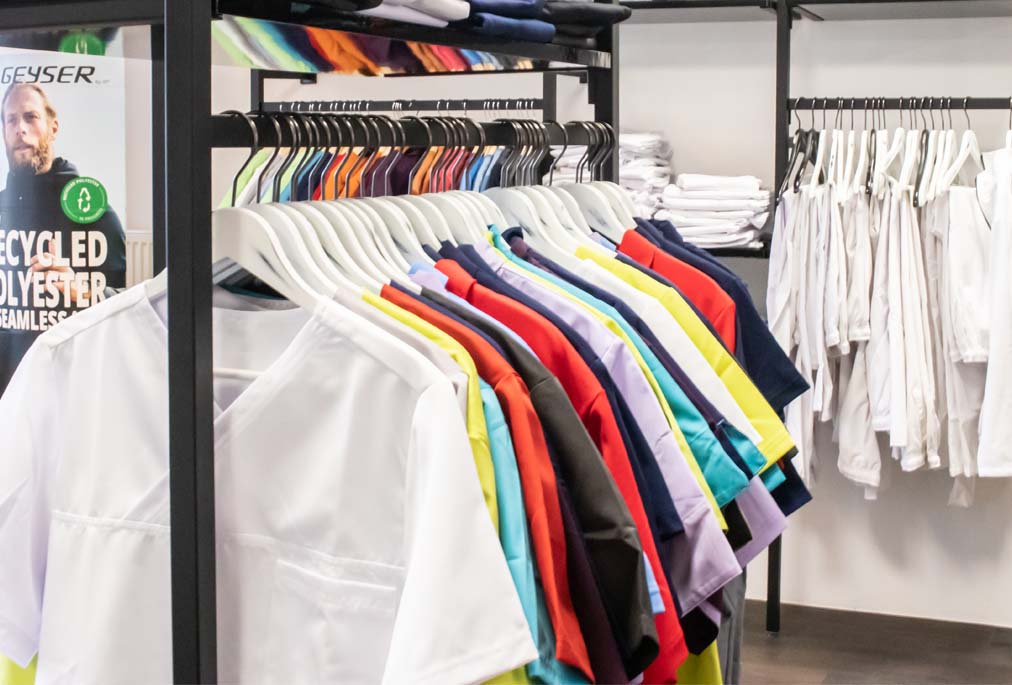 Bunte und weiße Pflegebekleidung für Damen und Herren auf Kleiderbügeln in einem Geschäft.