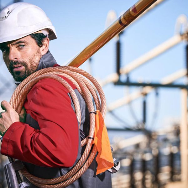 Elektriker in roter Störlichtbogen-Schutzkleidung und weißem Helm, trägt Kabel und Werkzeug.