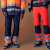Zwei Personen tragen Warnschutzbekleidung: orange-blaue Jacken und Hosen mit reflektierenden Streifen.